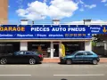 Garage avec enseigne bleue et jaune, deux voitures garées devant, sous un ciel bleu avec des nuages.