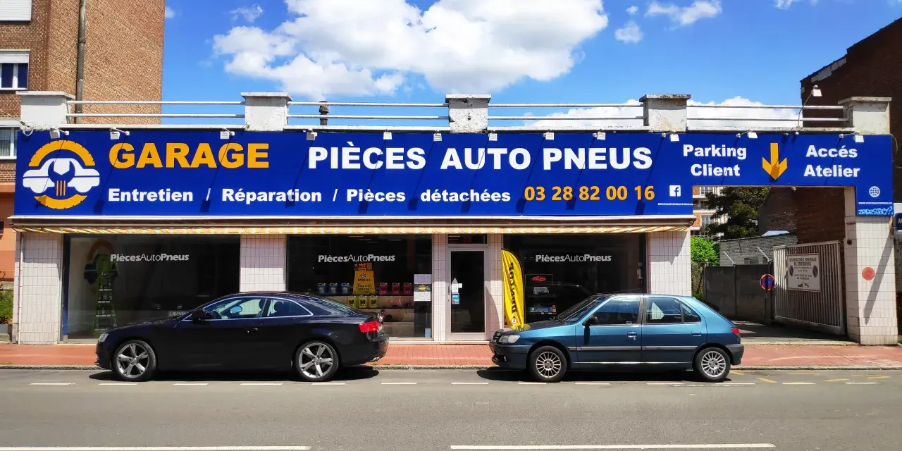 Garage avec enseigne bleue et jaune, deux voitures garées devant, sous un ciel bleu avec des nuages.