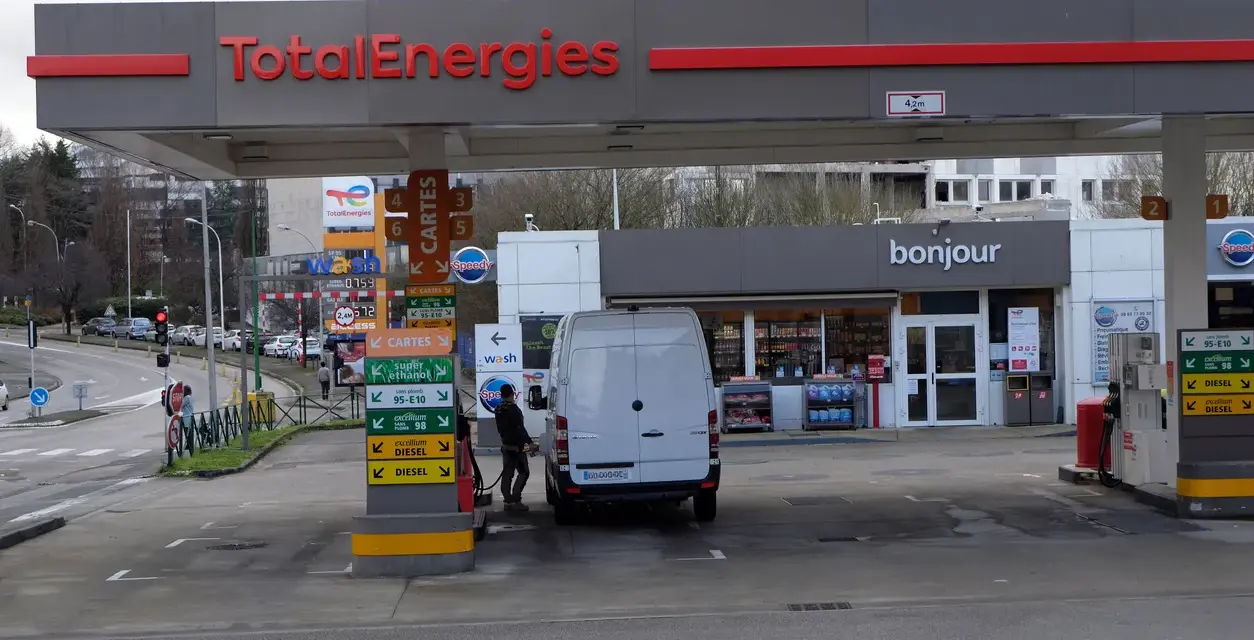 Station TotalEnergies avec une camionnette blanche en cours de ravitaillement. Panneaux de prix et de services.