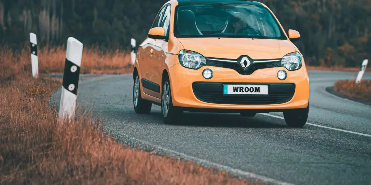 Une Twingo orange roule sur une route sinueuse bordée d'herbes sèches.