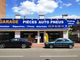 Garage avec enseigne bleue et jaune, deux voitures garées devant, sous un ciel bleu avec des nuages.