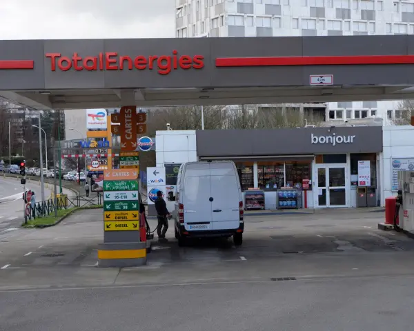 Station TotalEnergies avec une camionnette blanche en cours de ravitaillement. Panneaux de prix et de services.